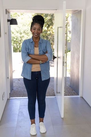 Smiling woman standing in open doorway at home welcoming in denim outfit and sneakers