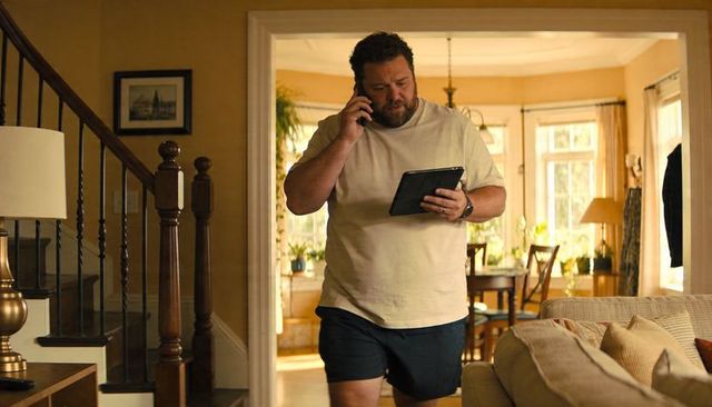 Bearded man walking through home talking on phone holding tablet multitasking in living room
