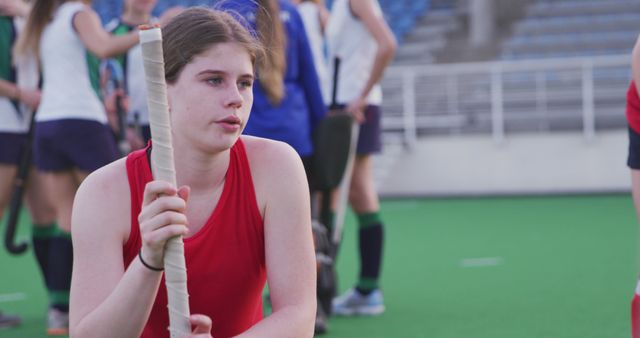 Young female field hockey player is taking a break during practice on a field. She is holding her hockey stick and appears focused. Field background with other players suggests a team training session. Ideal for sports magazines, athletic gear promotions, and articles on teamwork in sports.