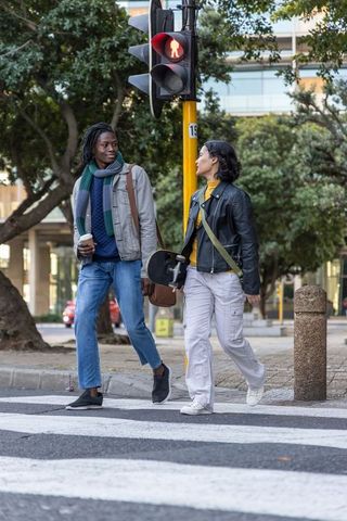 Young diverse friends walking across urban crosswalk carrying skateboard and coffee in casual style