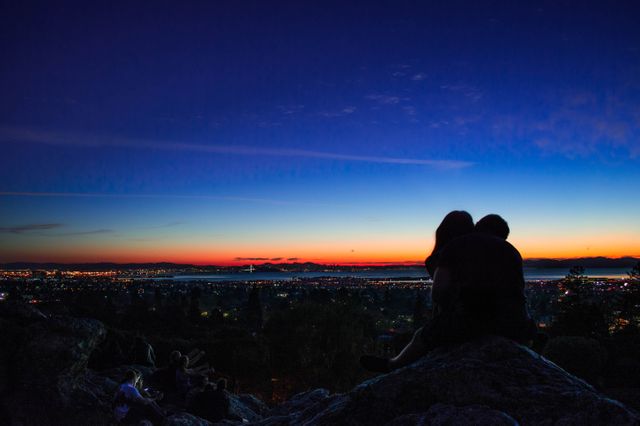 Silhouetted couple sitting close, enjoying colorful sunset over a vast cityscape view. Twilight transitioning with horizons glowing shades. Versatile for themes on love, romance, city living, or serene nature moments in urban settings. Perfect for marketing campaigns, advertisements, or nature and travel articles.