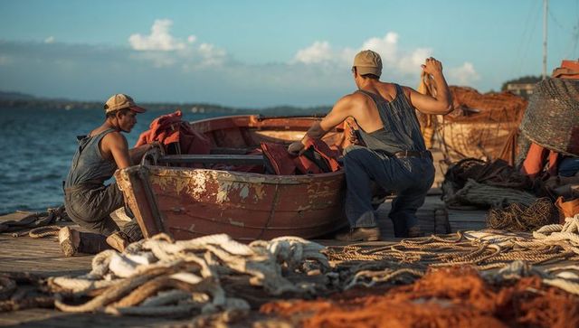 Two fishermen preparing weathered wooden skiff on harbor dock surrounded by ropes and nets