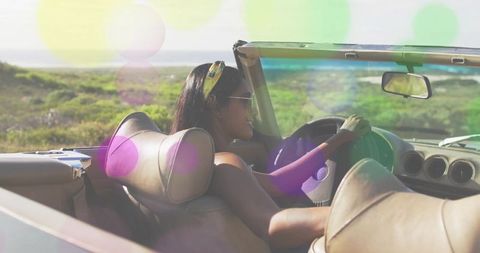 Woman driving convertible along coastal dunes wearing sunglasses and colorful headband