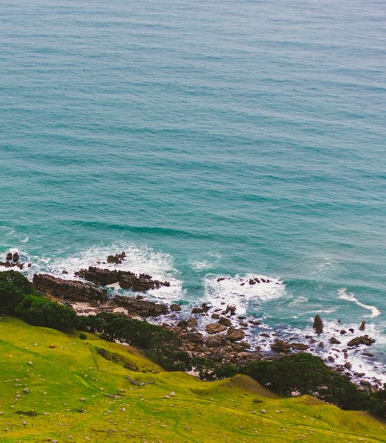 This coastal scene shows majestic green hills rolling down to meet the blue sea with waves crashing against a rocky shore. Suitable for use in travel magazines, nature articles, tourism brochures, or backgrounds in environmental blogs highlighting the beauty of nature and coastal landscapes.