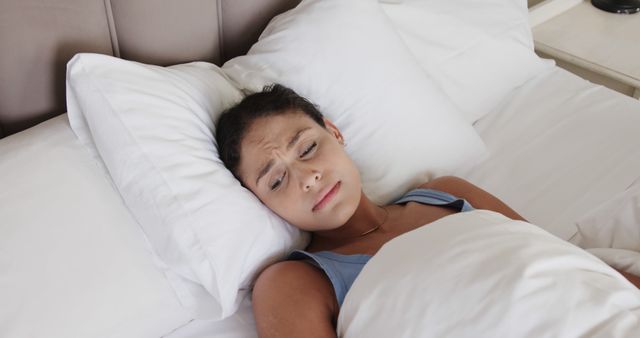 Young woman lying in bed appears restless and frustrated, suggesting difficulty sleeping. White bedding creates a clean and calming environment. Useful for illustrating topics on insomnia, sleep disorders, bedtime routines, and relaxation methods.