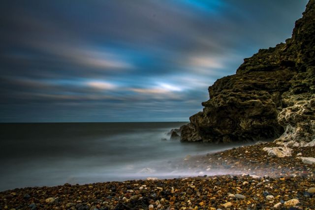 Pebbled beach with smooth waves created by long exposure under a cloudy sky at twilight. This tranquil coastal scene is perfect for use in landscape and nature photography, travel publications, and meditation or relaxation themes.