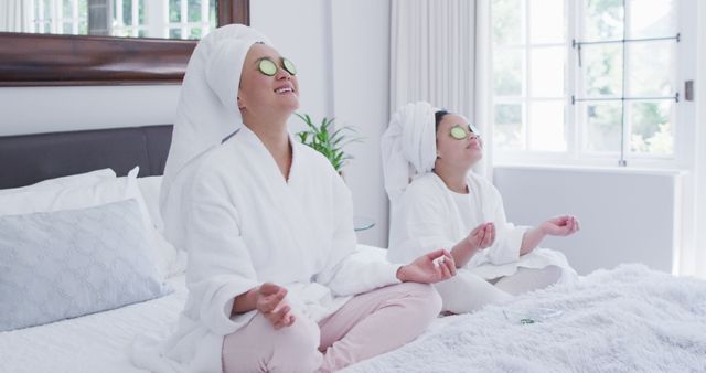 Mother and daughter wearing white robes and towels sitting on bed with cucumbers on eyes, enjoying a spa day at home. Both are sitting cross-legged, pottering around meditation and self-care activities. This can be used for promotions on family health, spa services, wellness blogs, and beauty treatment businesses.