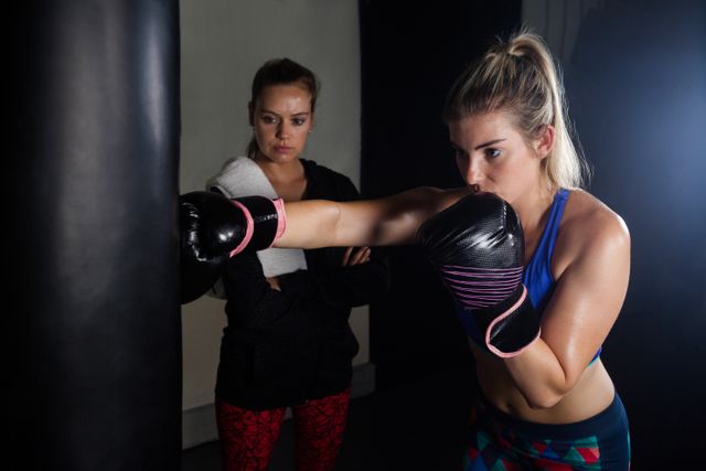 Woman intensely boxing while trainer observes and assists in a fitness studio. Ideal for promoting fitness training, personal coaching services, sports motivation, and athletic wear. Use in articles or advertisements focused on boxing training, fitness routines, and strength building.