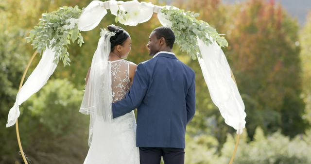 Bride and groom standing together under decorated wedding arbor, gazing into each other's eyes with love. Bridal gown features beautiful lace detail and long veil. Scene set outdoors with natural greenery and colorful foliage in background. Ideal for websites on wedding planning, bridal magazines, and articles about wedding trends and ceremonies.