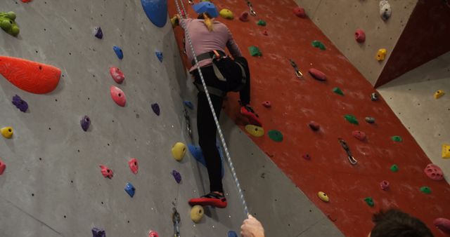 Woman climbing indoor rock wall using safety ropes, a setting perfect for illustrating adventure, fitness activity, and sports. Useful for promoting gyms, fitness centers, or adventure sport events. Can also be used in content about personal challenges, athletic training, and health benefits of rock climbing.