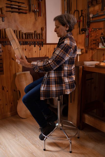 Side view of a senior Caucasian female luthier sitting and working on a violin at a workbench in her workshop, inspecting her handiwork and holding the unfinished violin body, with tools hanging up on the wall in the background.