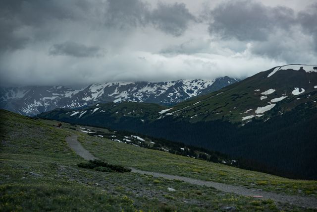 This captivating landscape shows a mountain range with patches of snow under dark, stormy clouds, with a rocky path leading through the wilderness. Ideal for use in nature magazines, outdoor adventure promotions, and travel brochures.