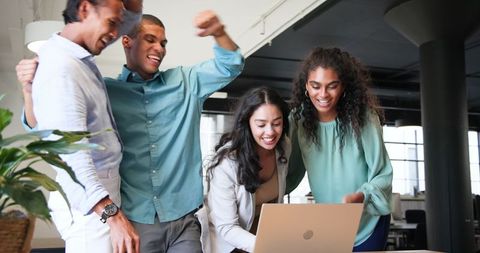 Diverse team celebrating success around laptop in modern office