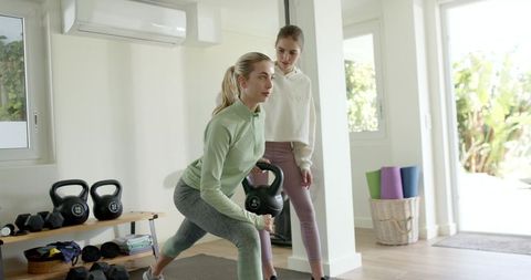 Woman performing kettlebell lunge while trainer coaching in sunlit home gym