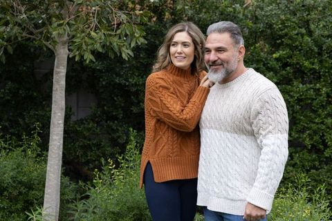 Mature couple wearing cozy cable-knit sweaters smiling while standing in leafy backyard
