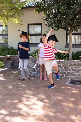 Energetic kids jumping with graduation caps and diplomas in sunny brick courtyard