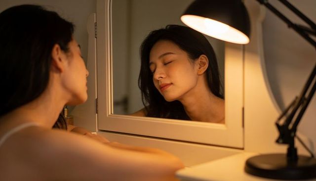 Asian woman relaxing at vanity mirror smiling with eyes closed in warm lamp light