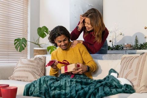 Diverse couple unwrapping gift on cozy living room sofa, festive holiday surprise moment