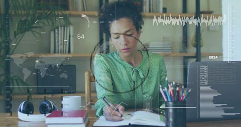 Mature Woman Working from Home Writing in Notebook at Desk with Laptop and Headphones