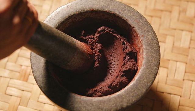 Stone mortar and pestle grinding reddish-brown spice paste on woven bamboo mat
