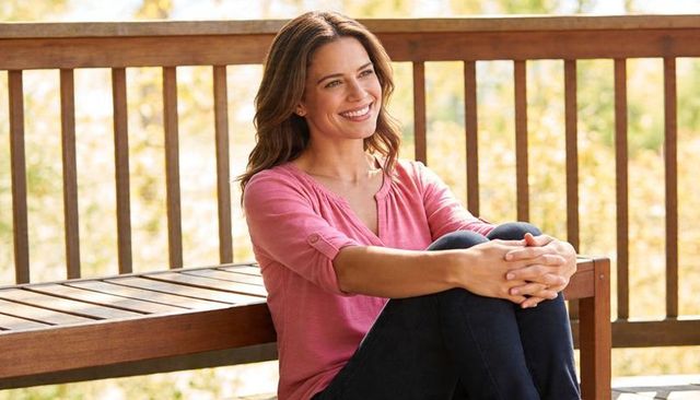 Sunlit woman sitting on porch bench hugging knees, smiling, wearing pink henley and jeans