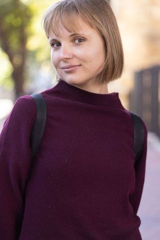 Young woman in maroon sweater smiling on sunlit city sidewalk with backpack straps