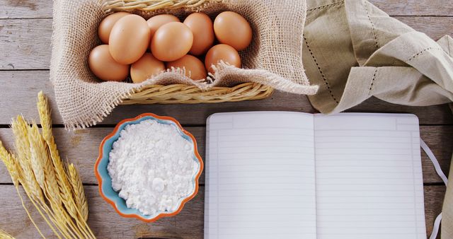 A variety of baking ingredients are laid out on a wooden surface, including eggs, flour, and wheat, alongside an open notebook with copy space. It suggests preparation for baking, with the notebook used for jotting down recipes or notes.