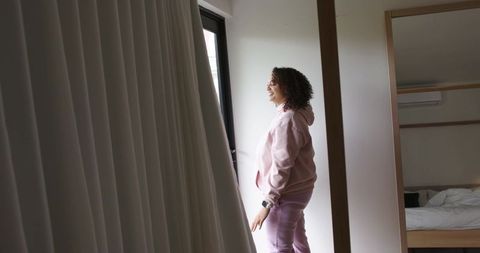 African american woman standing by window in modern bedroom in pink loungewear checking smartwatch