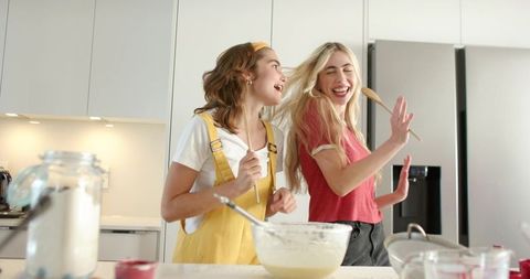 Two friends singing and baking at kitchen island while mixing batter with wooden spoon