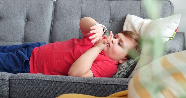 A young boy in a red shirt is lying on a gray couch, engrossed in a handheld device. This calm and casual scene is perfect for illustrating childhood leisure activities at home. It could also be used in discussions about the influence of technology on children, or in promotional materials for family-oriented products.