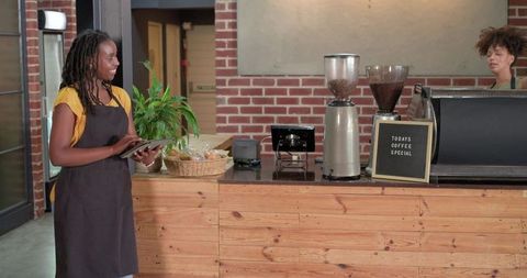 Barista holding tablet coaching coworker preparing espresso at rustic wood coffee counter