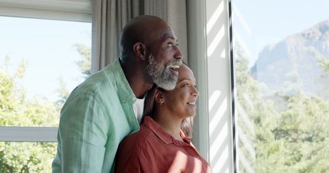 Mature couple standing by window gazing at mountain vista, sunlit peaceful intimate moment
