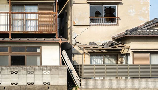 Residential facade showing structural damage: cracked stucco, shattered window, collapsed roof