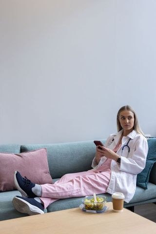 Clinician reclining on teal sofa during break, using smartphone, wearing scrubs and stethoscope