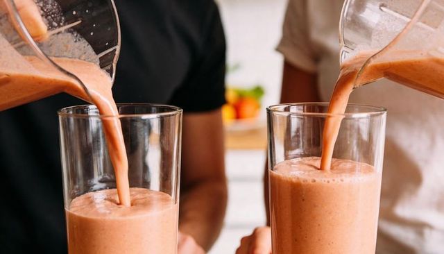 Pouring peach smoothie into two tall glasses with pitchers at home kitchen counter