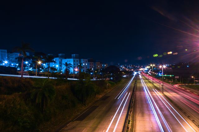 Captures vibrant night city scene with light trails from vehicles on highway, creating motion blur. Urban landscape features illuminated streetlights and modern buildings. Useful for illustrating busy city life at night, high-speed traffic, and concepts of transportation and energy.