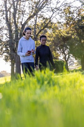 Multicultural couple walking through sunlit park meadow while checking smartphone together