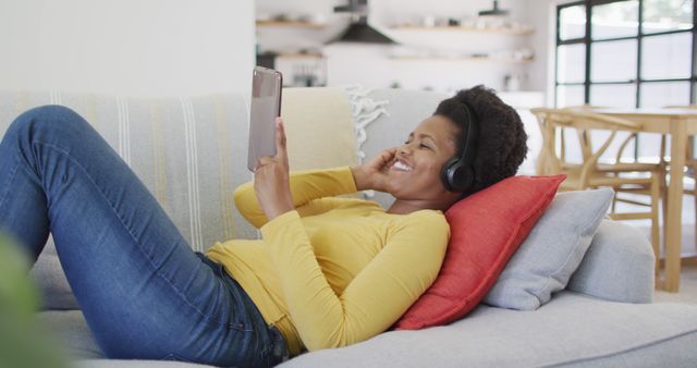 Smiling woman laying on couch, using a digital tablet and wearing headphones in a casual indoor setting. Perfect for promoting technology products, leisure activities, or home-related services. Ideal for lifestyle blogs, advertisements, and articles focusing on relaxation, happiness, or modern living.