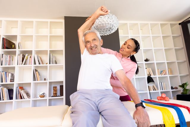 A biracial female physiotherapist is stretching a senior man's arm during a home exercise session. The setting is a well-lit room with a bookshelf in the background, indicating a comfortable home environment. This image can be used for articles or advertisements related to physical therapy, senior healthcare, home rehabilitation, and wellness programs for the elderly.