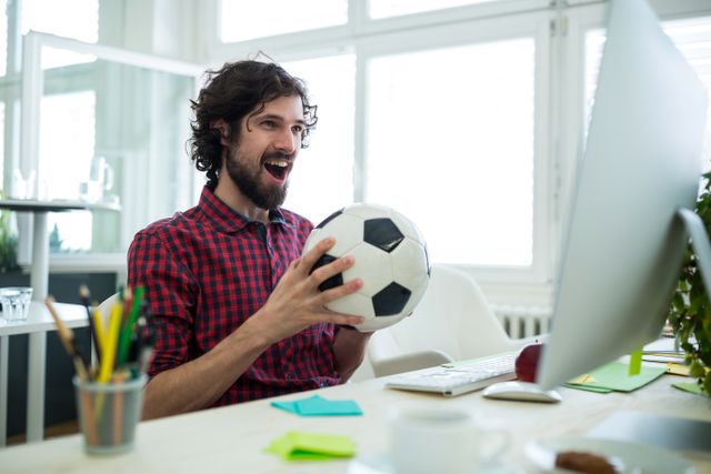 Businessman holding soccer ball, watching football match on computer in modern office. Ideal for illustrating sports enthusiasm in workplace, remote work culture, or balancing work and leisure.