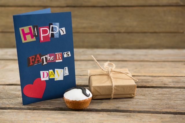 Father's Day greeting card with a heart design, a small gift box tied with twine, and a cupcake with a mustache decoration on a wooden table. Ideal for use in holiday promotions, greeting card designs, and social media posts celebrating Father's Day.