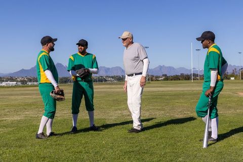 Veteran coach guiding baseball team during outfield drill on sunny field, bat gloves