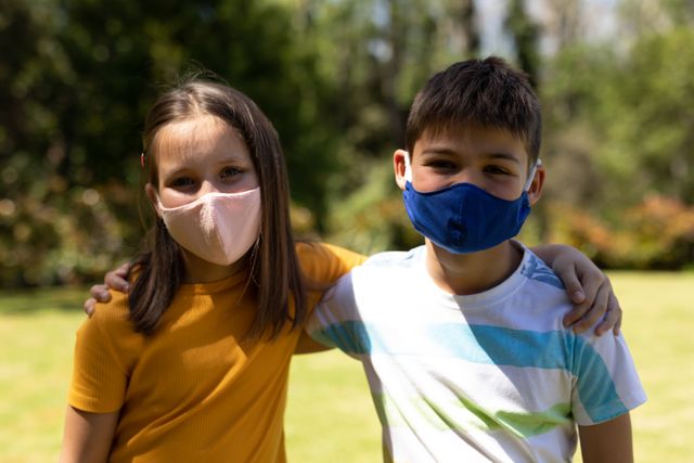 A caucasian boy and a girl wearing facemasks outside on a bright and sunny day. both of them have their arms on each other's shoulders.