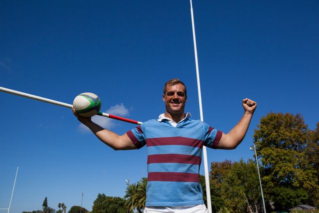 Portrait of happy rugby player holding ball with arms raised by goal post against blue sky