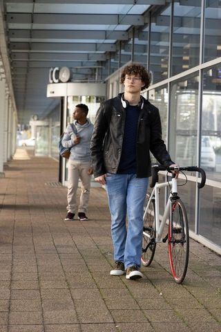 Young man walking bicycle beside glass facade on covered city walkway, commuter lifestyle