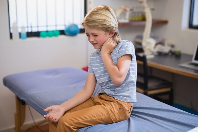 Young boy sitting on examination table in medical clinic, holding neck in pain. Ideal for use in healthcare, pediatric care, medical treatment, and injury-related content. Can be used in articles, blogs, and educational materials about children's health, injury prevention, and medical consultations.