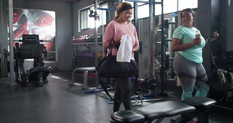 Diverse women walking and talking in gym weights area carrying duffel bag and shaker