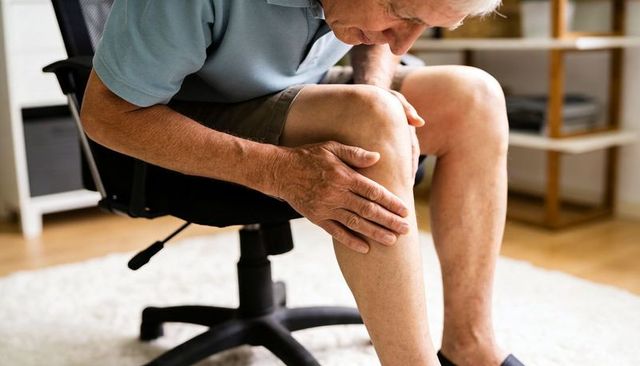 Senior man holding painful knee while sitting on swivel chair at home showing leg pain