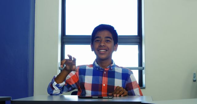 Boy wearing checked shirt, sitting in front of a large window. Ideal for educational technology, learning activities, classroom environment and school-related content.