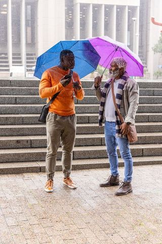 African american men talking under colorful umbrellas checking phone on rainy urban steps